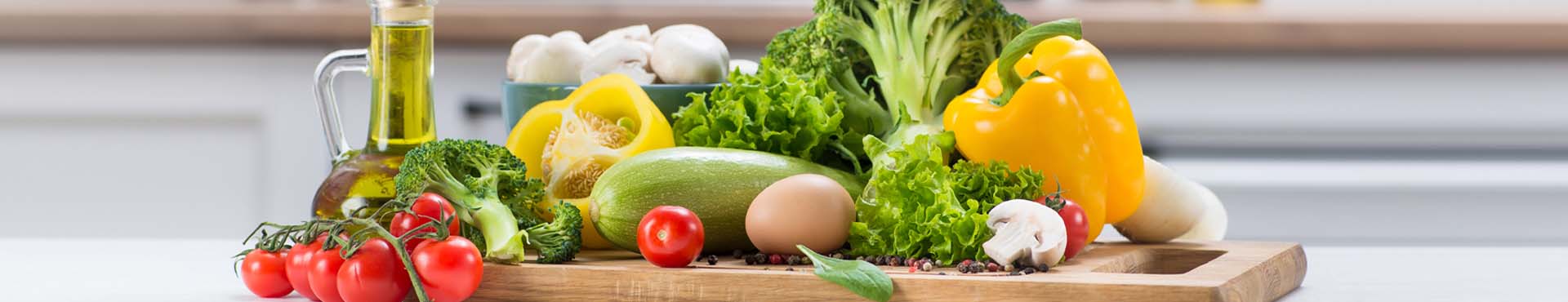 Aufnahme von gesunden Lebensmitteln, wie Salat, Brokkoli, Tomaten, Pilzen, Olivenöl - arrangiert auf einem Holzbrett vor dem unscharfen Hintergrund einer Küche